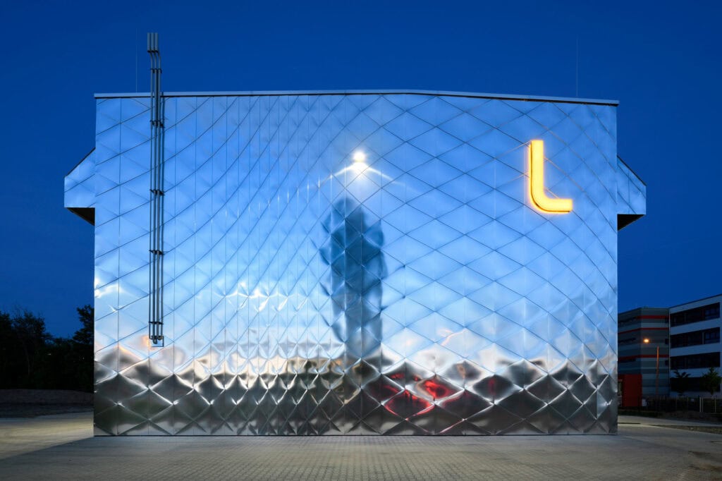 Eine silber strahlende, vorgehängte hinterlüftete Fassade an einem Blockheizkraftwerk nähe Leipzig. Ein orange leuchtendes "L" zieht das Gebäude bei tollem Licht.